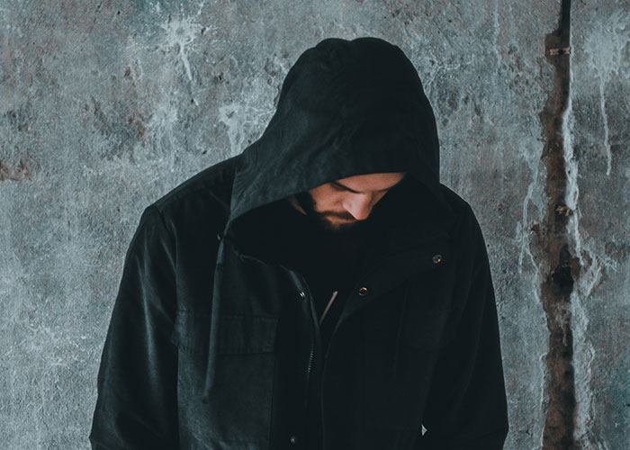 Man in a black hooded jacket looking down against a concrete wall, representing people living double lives and deception.