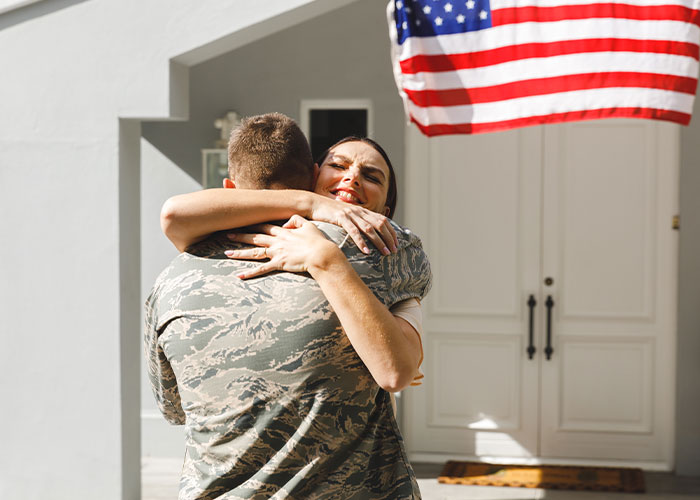 Person in military uniform hugging a smiling woman outside a house with an American flag, hinting at living double lives.
