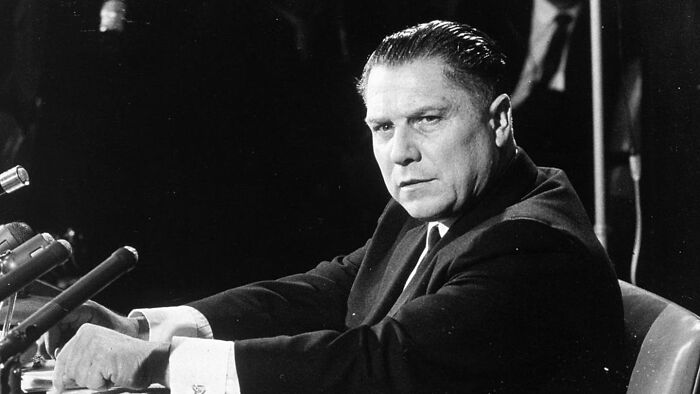Black and white image of a serious male celebrity speaking at a press conference about vanished celebrities and conspiracy theories.