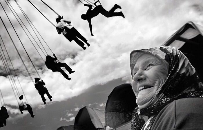 Elderly woman smiling in a street setting with people swinging on a carousel, capturing the beauty and wisdom of old age.