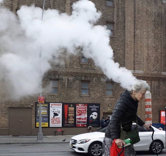 Elderly woman with gray hair walking on street near a steam vent, capturing beauty and wisdom of old age in a street photo.