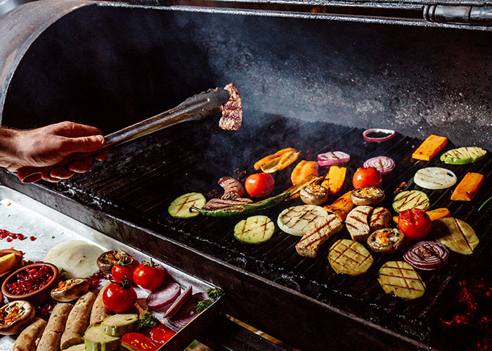 Grilling assorted vegetables and meats with tongs over a smoky open flame, food industry workers handling customer orders carefully.