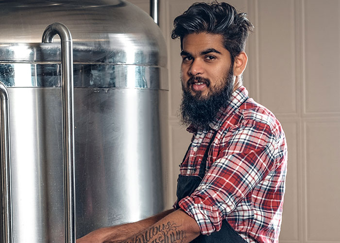 Bearded food industry worker in plaid shirt standing next to large stainless steel brewing equipment inside brewery.