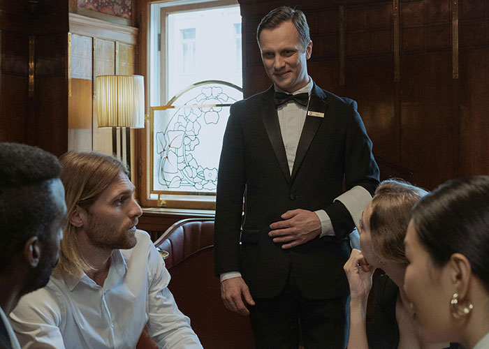 Waiter in a formal suit attending to a group of restaurant customers in a vintage-style dining room setting.