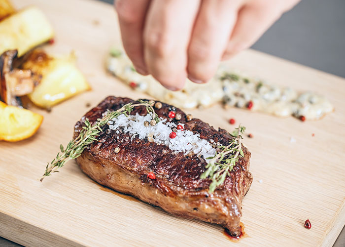 Close-up of a seasoned steak on a wooden board with herbs and pepper, illustrating dumbest customers in the food industry.