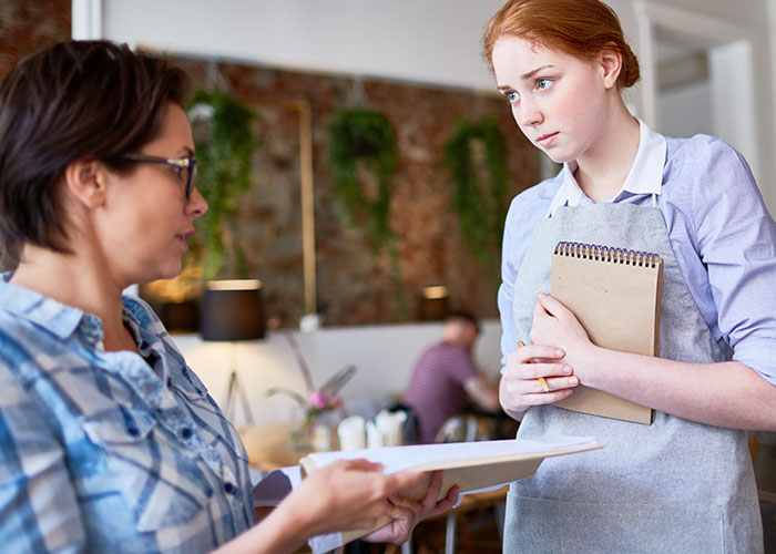 Customer speaking to a serious waitress holding a notepad in a restaurant, illustrating dumbest customers in food industry.