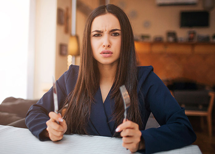 Confused young woman holding fork and knife at restaurant table, representing dumbest customers in food industry stories.