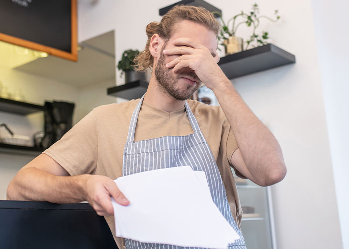 Food industry worker in apron facepalming while holding papers, overwhelmed by dumbest customers experiences.