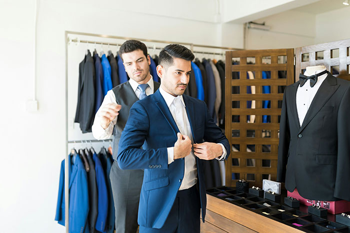 Man trying on a blue suit in a store while another man assists, illustrating unhinged company credit card purchases.