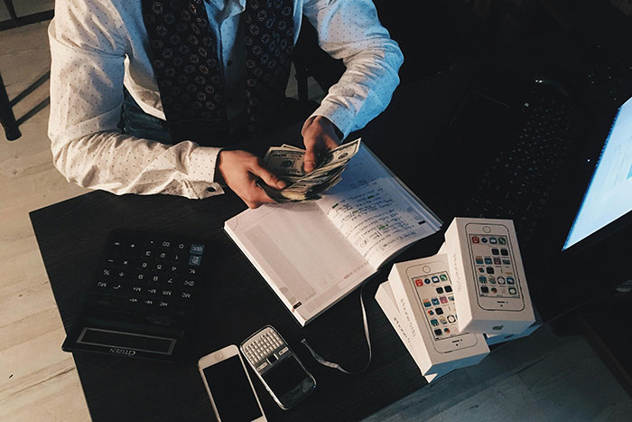 Person counting cash at a desk with multiple new smartphone boxes and electronic devices representing company credit card use.