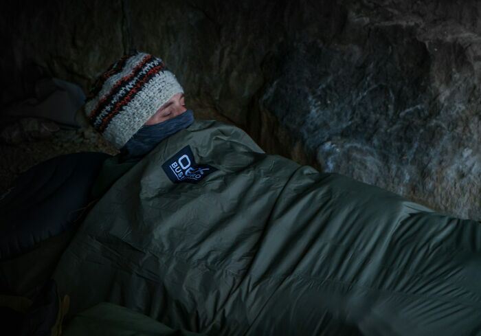 Person wrapped in a sleeping bag inside a dark cave, evoking the eerie atmosphere of creepy stories in the woods.