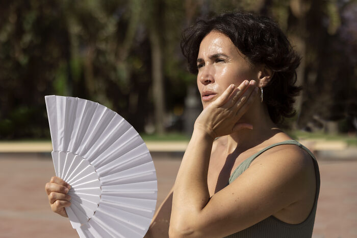 Woman using a hand fan outdoors looking uncomfortable and overheated, illustrating advanced stupid out-of-touch behavior.