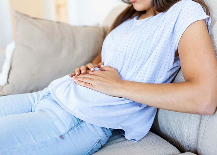 Woman in casual clothes sitting on a couch holding her stomach, illustrating facepalm-worthy doctor’s visits.