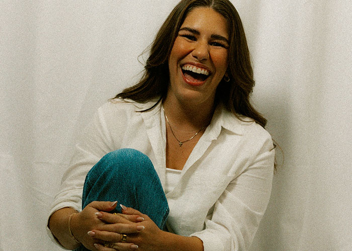 Young woman laughing joyfully, wearing a white shirt and jeans, expressing hilariously embarrassing doctor’s appointment moments.
