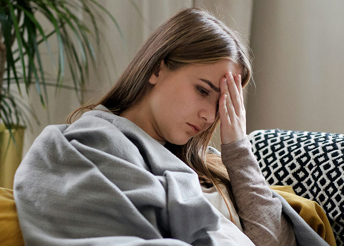 Young woman wrapped in a blanket sitting on a couch, looking stressed and frustrated after a doctor’s visit.