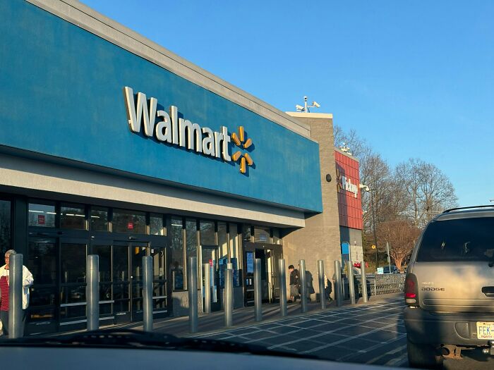 Exterior view of Walmart store entrance with cars in parking lot and people walking nearby during daytime.
