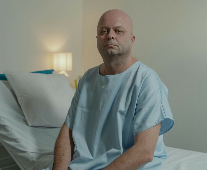 Bald man in hospital gown sitting on bed, representing cancer diagnosis and betrayal from hidden truth.