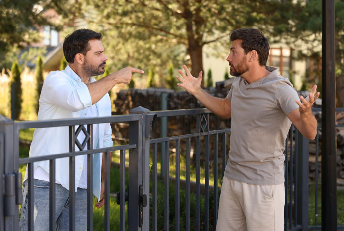 Two entitled neighbors arguing loudly over a blocked driveway next to a metal fence in a residential area.
