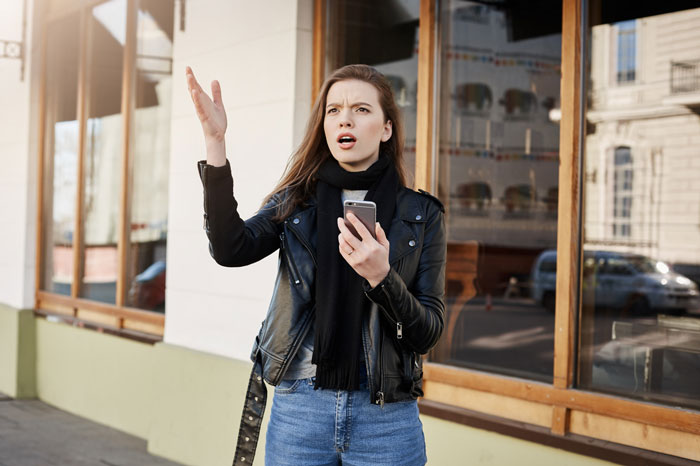 Frustrated young woman holding phone outside, upset about entitled neighbors blocking driveway on a city street.