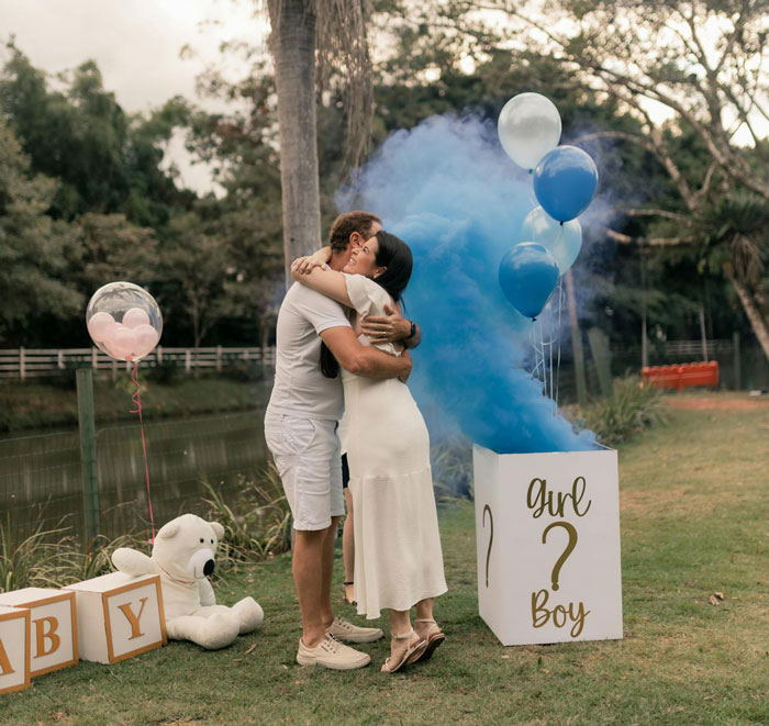 Couple hugging near gender reveal box with blue smoke and balloons, related to sister adoption papers after babysitting mix-up.