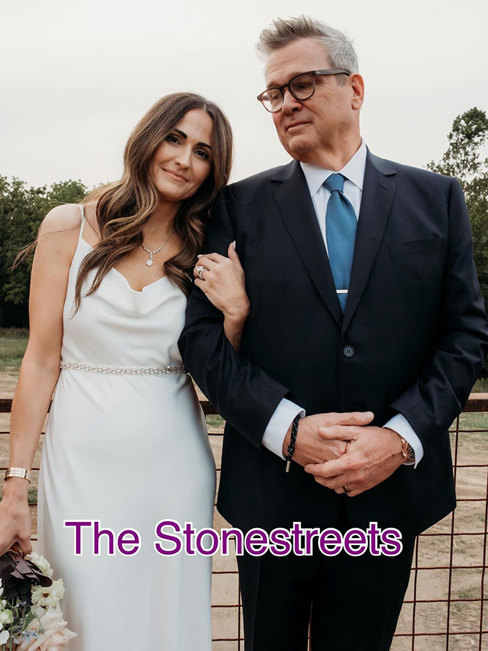 Eric Stonestreet in a dark suit with a woman in a white dress, celebrating their wedding day outdoors.