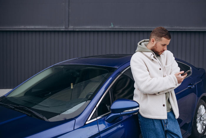 Young man leaning on blue car, checking phone outside a garage, depicting entitled neighbor trying to hijack driveway.
