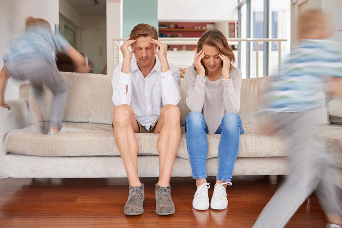 Frustrated parents sitting on a couch with blurred children running around, stressed about stepdaughter dating and love nest.