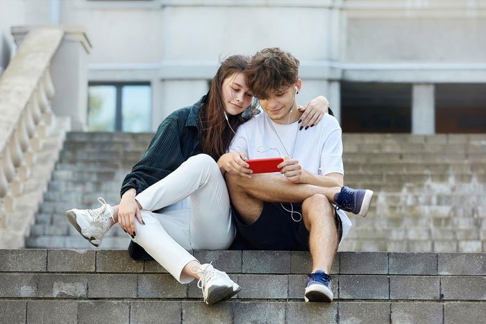 Teen couple sitting on steps enjoying a video on a red phone, highlighting stepdaughter dating against mom&rsquo;s wishes.