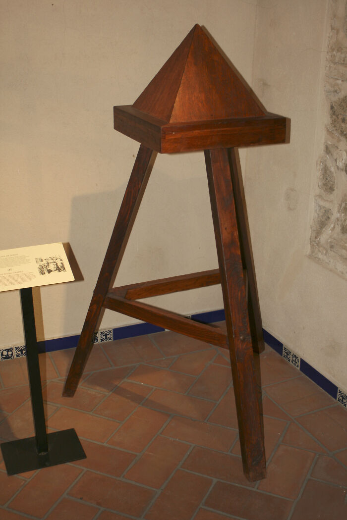 Medieval t*****e device with a wooden pyramid spike on three legs displayed on a tiled floor against a wall.