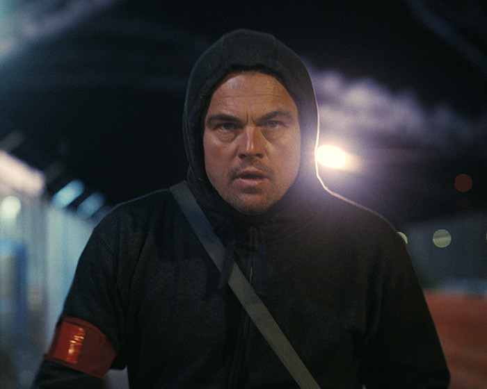 Leonardo DiCaprio in a dark hoodie running outdoors at night with an intense expression and steam in the background.