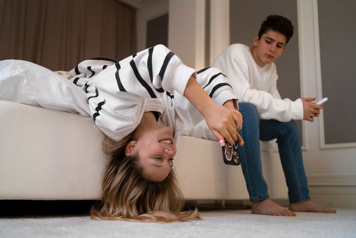 Two siblings in a bedroom, a girl playing with her phone upside down while her brother watches, reflecting child support dynamics.