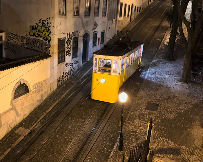 Yellow Lisbon tram traveling at night on cobblestone street near buildings and graffiti in urban setting.