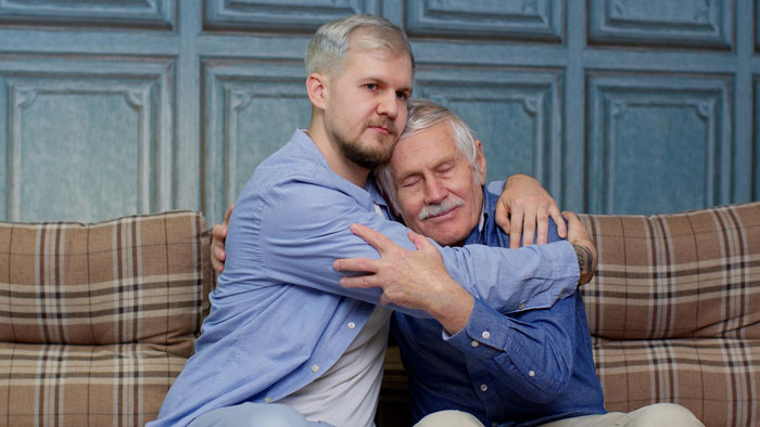 Two men hugging on a plaid couch, representing conflict over house ownership and relationship ultimatums. Two men hugging on a plaid couch, representing conflict over house ownership and relationship ultimatums.