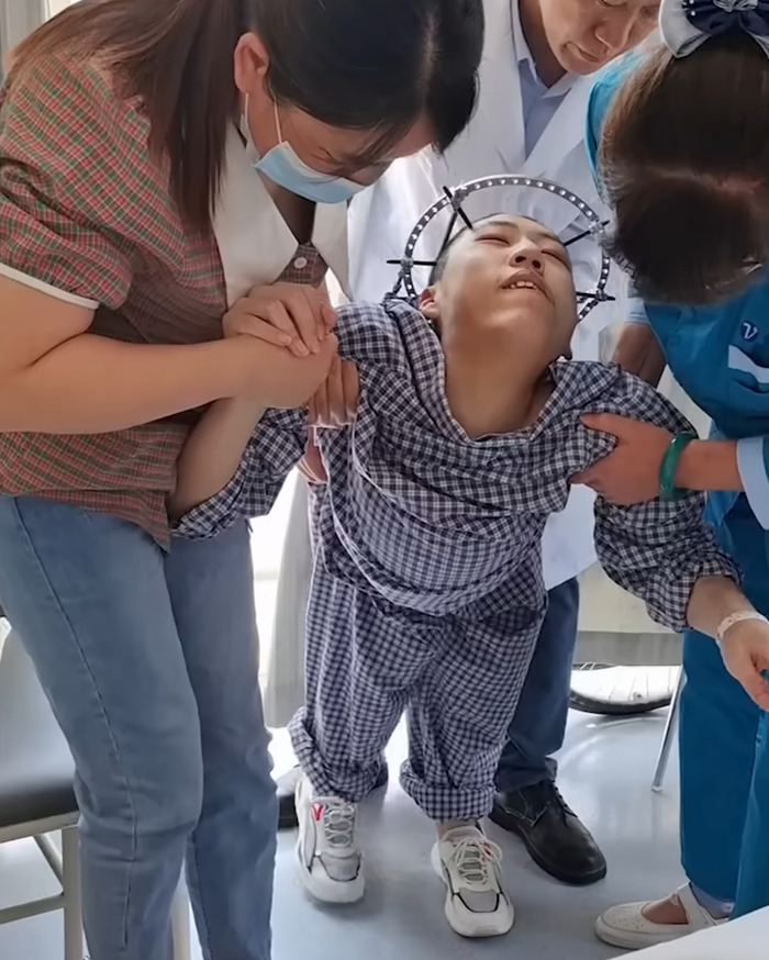 Young folded boy with head brace being supported by medical staff and a woman as he attempts to stand upright.