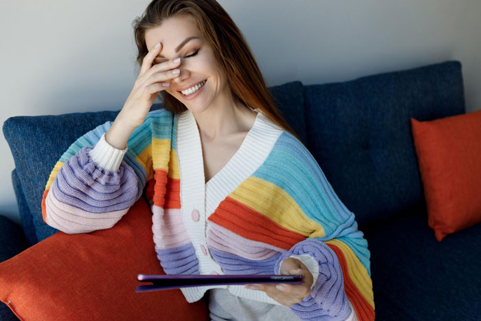 Young woman smiling and holding a tablet, sitting on a couch with colorful cushions and wearing a striped sweater.
