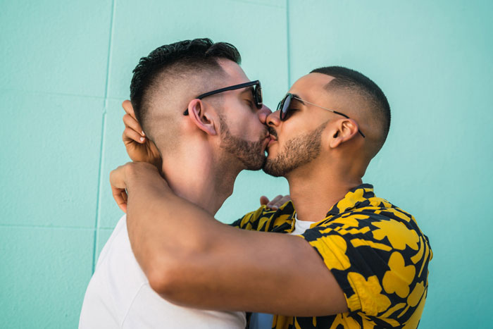 Groom kisses best friend at bachelor party in a close embrace, showing affection against a teal wall background.