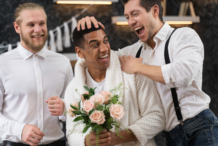 Groom laughing with best friends at bachelor party, holding bouquet, while celebrating before wedding day event.