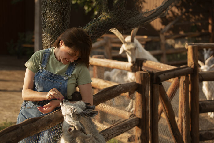 Woman in denim overalls feeding a goat at a farm, capturing an out-of-touch moment with animals and nature.