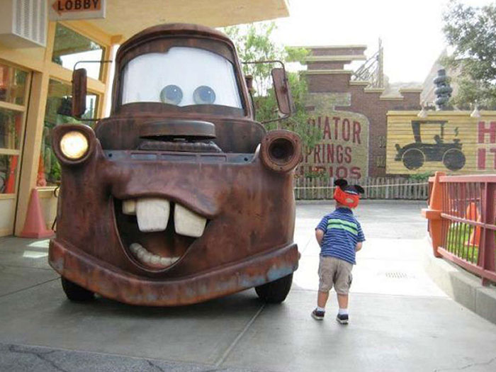 Boy wearing a cap standing near a large character car at an amusement park during a family trip.