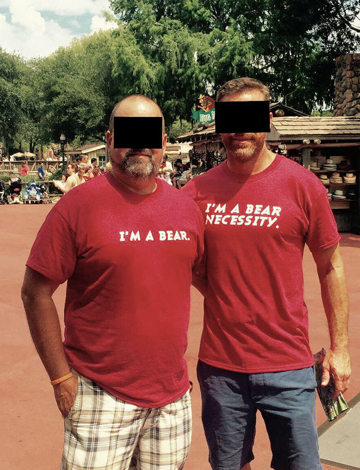 Two men wearing matching red shirts with bear-themed text at an amusement park during a daytime visit.