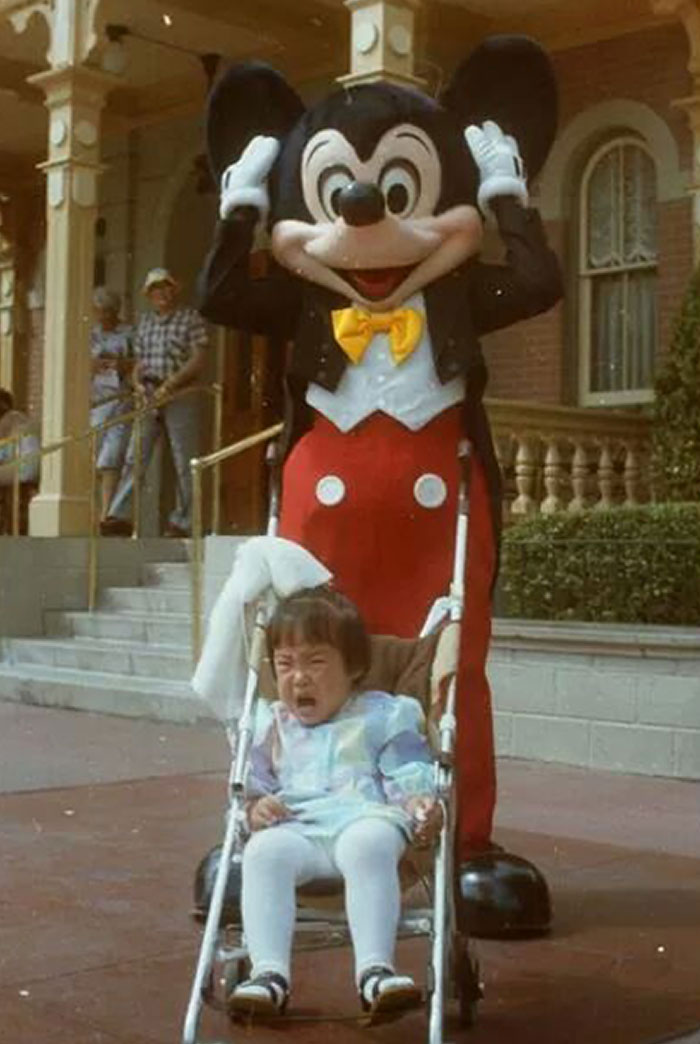 Child crying in stroller while posing with amusement park character mascot in a crowded park setting.