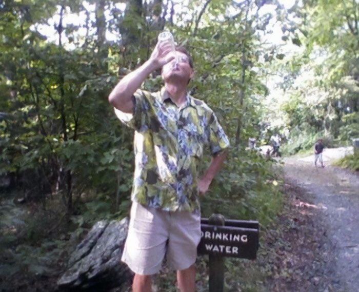 Man wearing floral shirt drinking water from a bottle near a drinking water sign in a forest, funny parents moment.