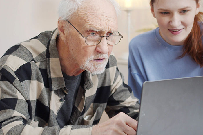 Older man struggling with technology while young woman helps him use a laptop illustrating clueless older people online.
