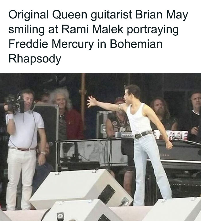 Brian May smiling at Rami Malek portraying Freddie Mercury during a live performance moment from Bohemian Rhapsody scene.