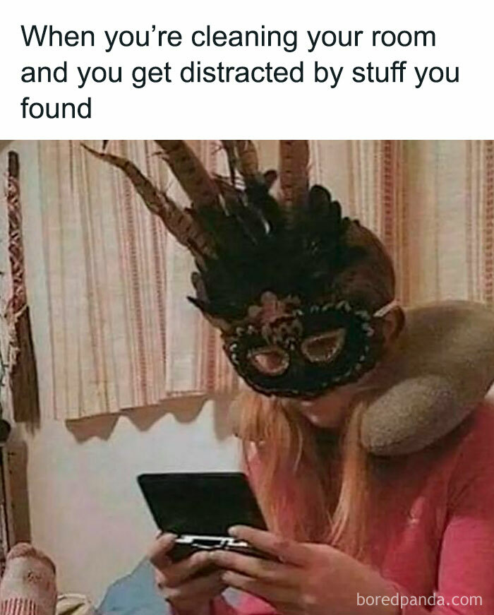 Person wearing a feathered mask and neck pillow, distracted by handheld gaming device, funny meme about cleaning room distraction.