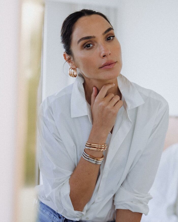 Gal Gadot wearing white shirt and gold bracelets, posing thoughtfully amid Venice Film Festival ban petition controversy.