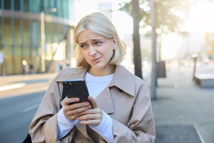 Woman outdoors looking confused while holding smartphone, illustrating entitled neighbor conflict and towing dispute.
