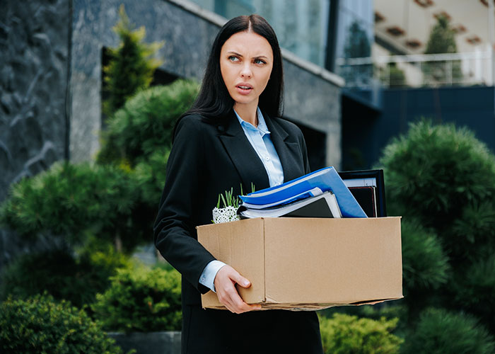 Woman employee carrying a box of office items outside a building, looking concerned after getting laid off.