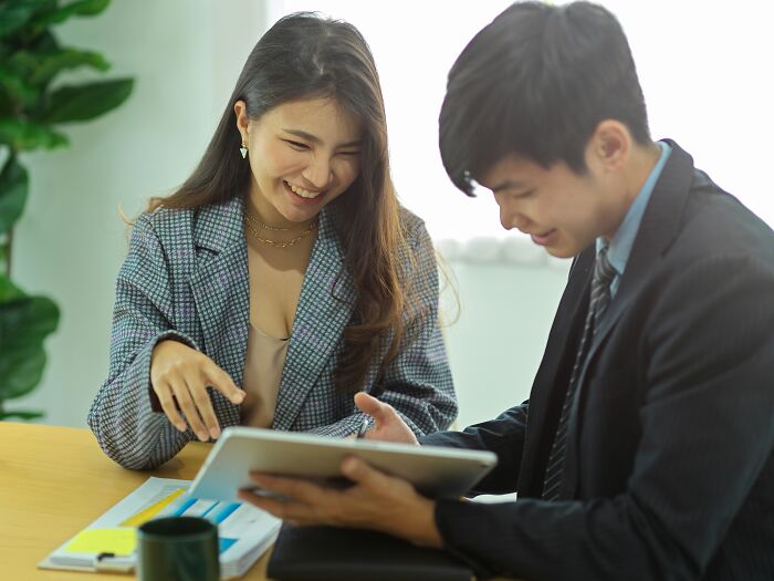 Two young professionals sharing things they would never admit in real life while discussing documents and tablet at office.