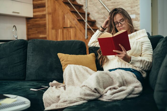Woman wrapped in blanket on sofa reading a book, reflecting on groom insisting guest attend wedding despite positive Covid test.